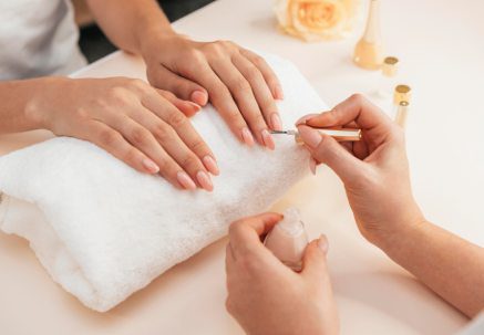 Observamos primer plano de manos tanto de cliente como esteticien. La cliente tiene sus manos sobre una toalla doblada y vemos que la esteticien está realizando una manicura. 