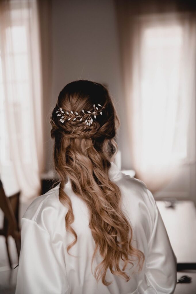 Novia de boda de espaldas frente a ventanas con cortinas donde podemos apreciar un precioso cabello largo recogido y con diadema floral. 